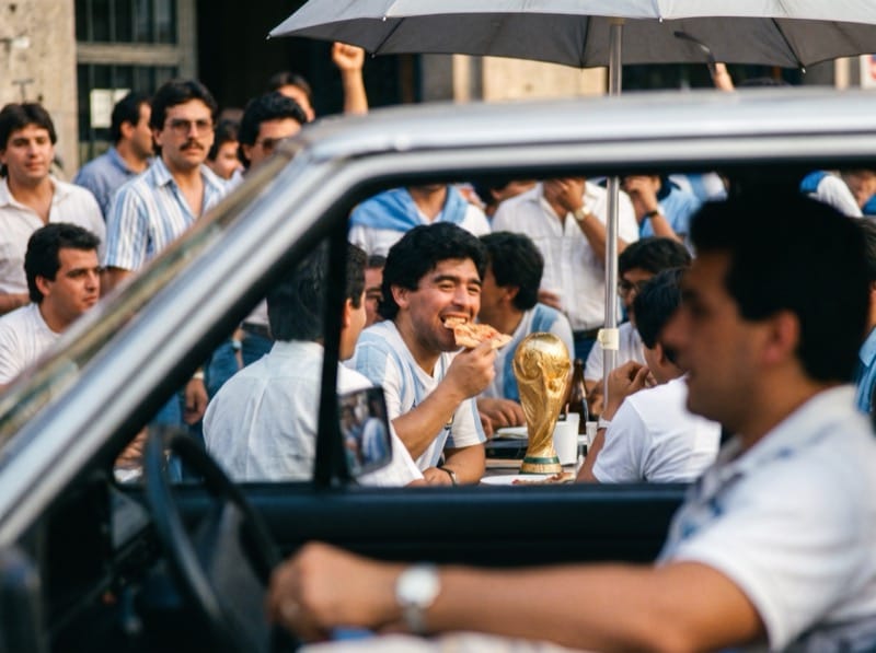 Diego entre la gente con una copa, visto desde un auto